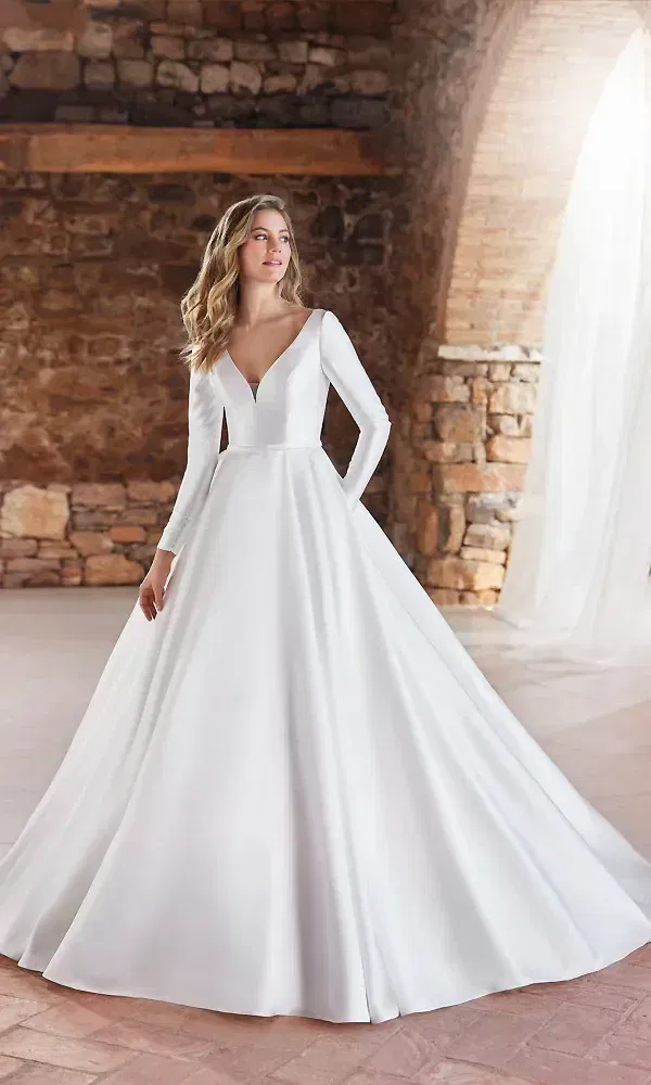 Schlichtes, aber luxuriöses Hochzeitskleid mit langen Ärmeln und einem weiten Rock. Das Kleid zeichnet sich durch seinen tiefen V-Ausschnitt und eine moderne, minimalistische Silhouette aus, perfekt für eine stilvolle Braut, die klassische Eleganz schätzt.