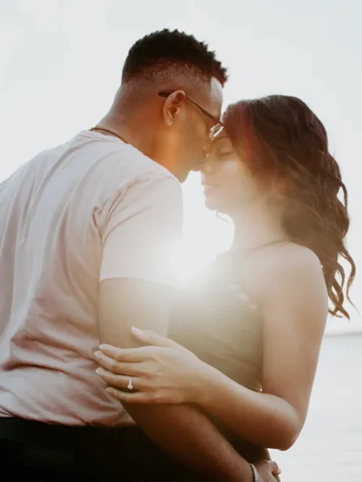 Ein Paar in einer innigen Umarmung vor einem strahlenden Sonnenuntergang. Ihre Stirnen berühren sich zärtlich, während die Frau ihre Augen geschlossen hält. Ihre Hand ruht auf seinem Arm und zeigt einen glänzenden Verlobungsring, der im Licht des Sonnenuntergangs funkelt. Das Bild vermittelt einen ruhigen und romantischen Moment.