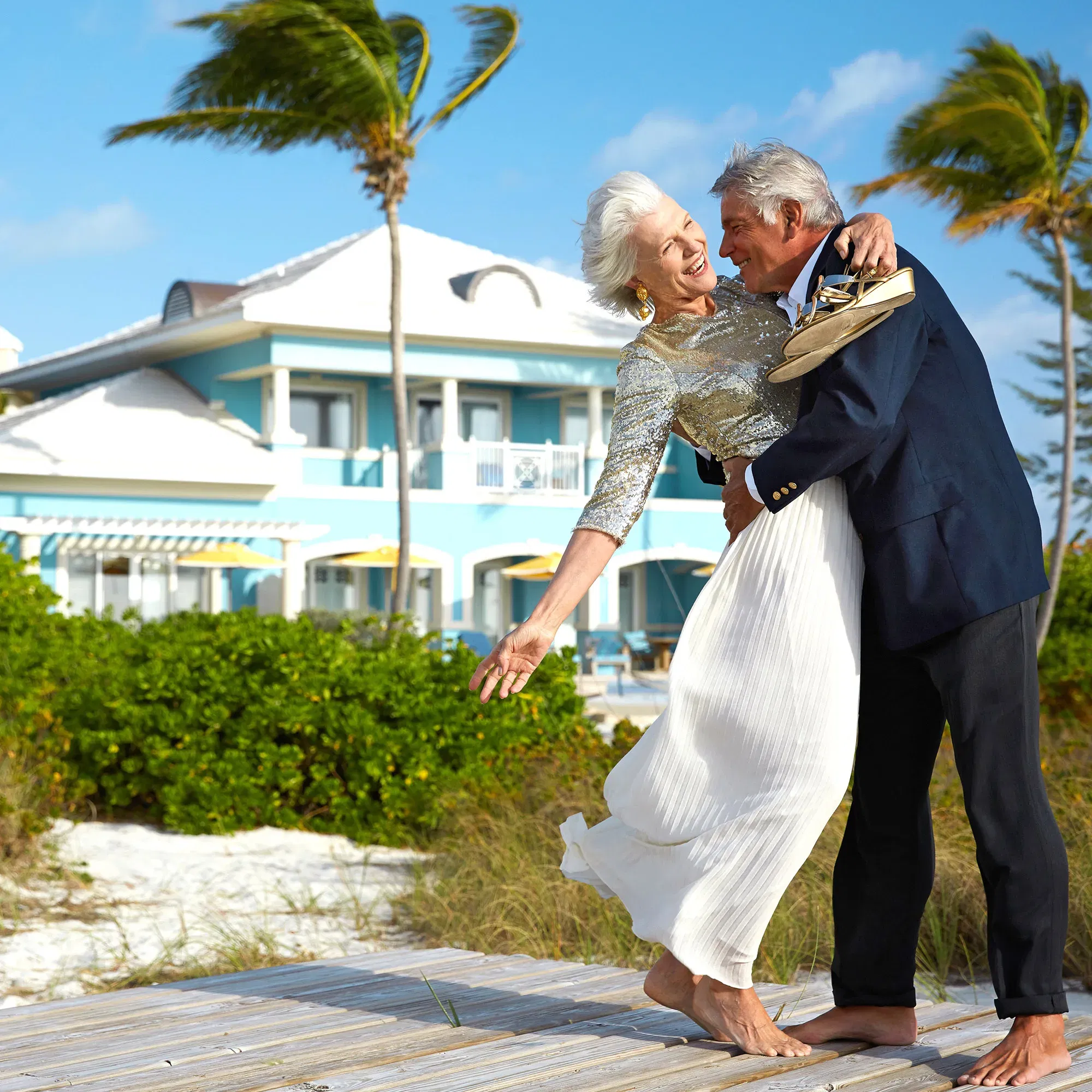 Ein älteres Paar in eleganter Kleidung, lachend und tanzend am Strand, während die Meeresbrise durch die Palmen weht.