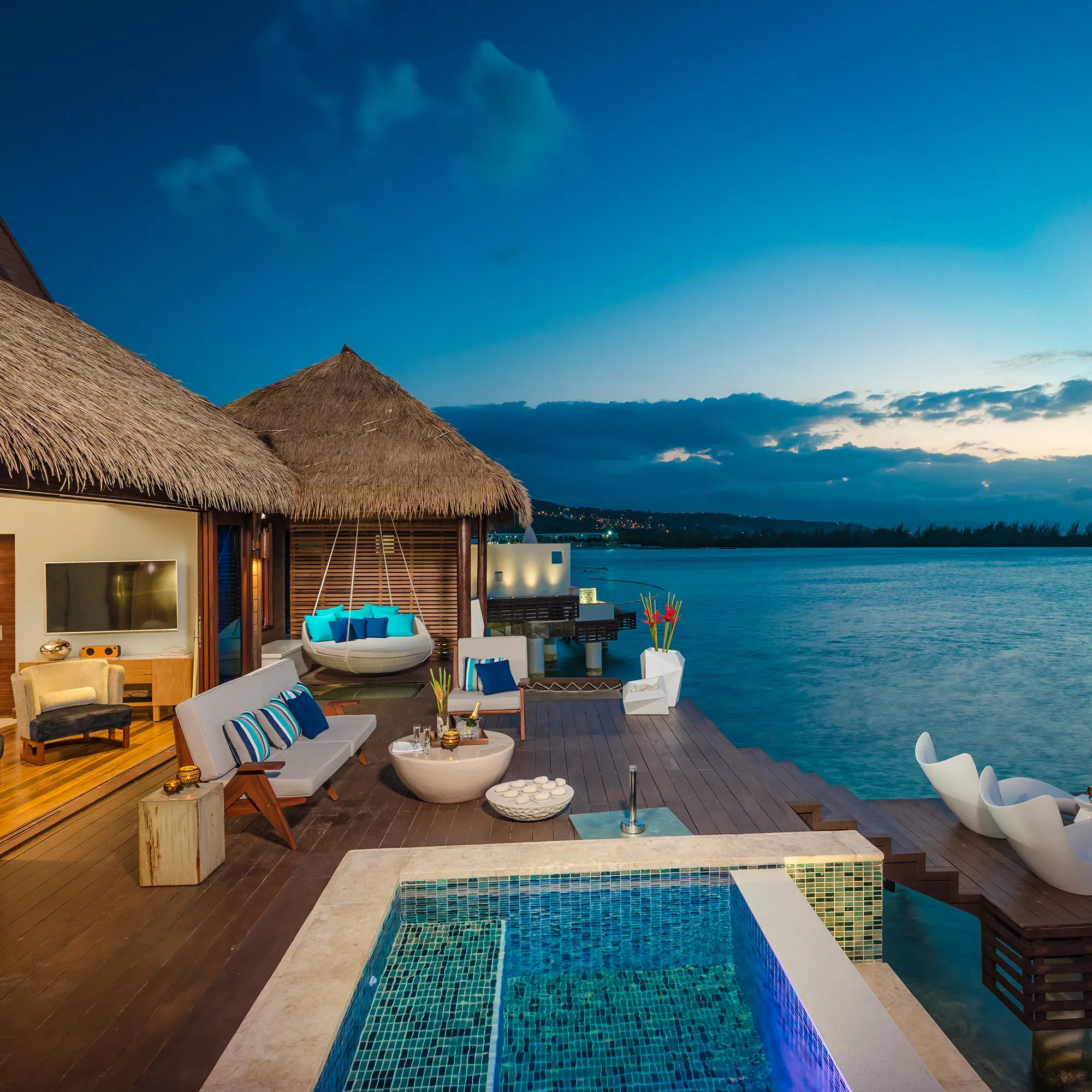 Ein tropischer Überwasser-Bungalow mit Pool, umgeben von Meer, stilvoll eingerichtet mit Hängesessel und Sitzgelegenheiten auf einer Terrasse bei Dämmerung.