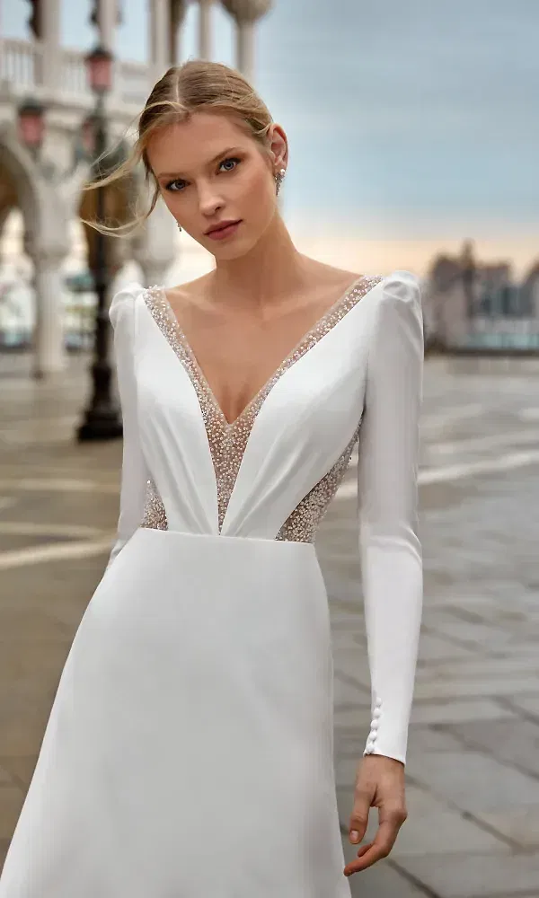 Eine Braut in einem schlichten, aber luxuriösen Hochzeitskleid mit einem funkelnden, tiefen V-Ausschnitt. Das Kleid besticht durch eine elegante Silhouette und langärmelige Raffinesse, während der Hintergrund eine architektonische Kulisse bietet.