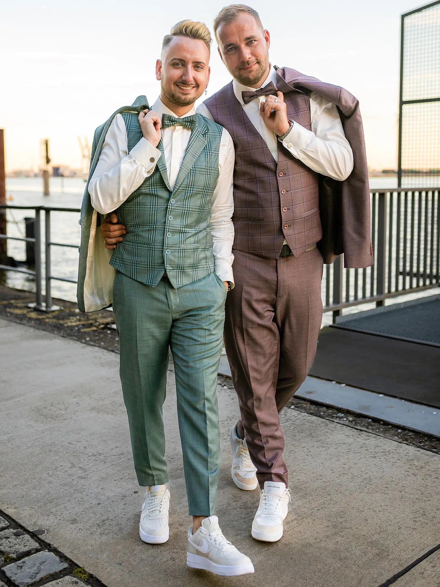 Zwei Männer posieren vor einem Hafen, beide in auffälligen Anzügen – einer in grün kariert, der andere in Lila. Sie tragen ihre Jacken über die Schultern und kombinieren den Look mit sportlichen weißen Sneakern.