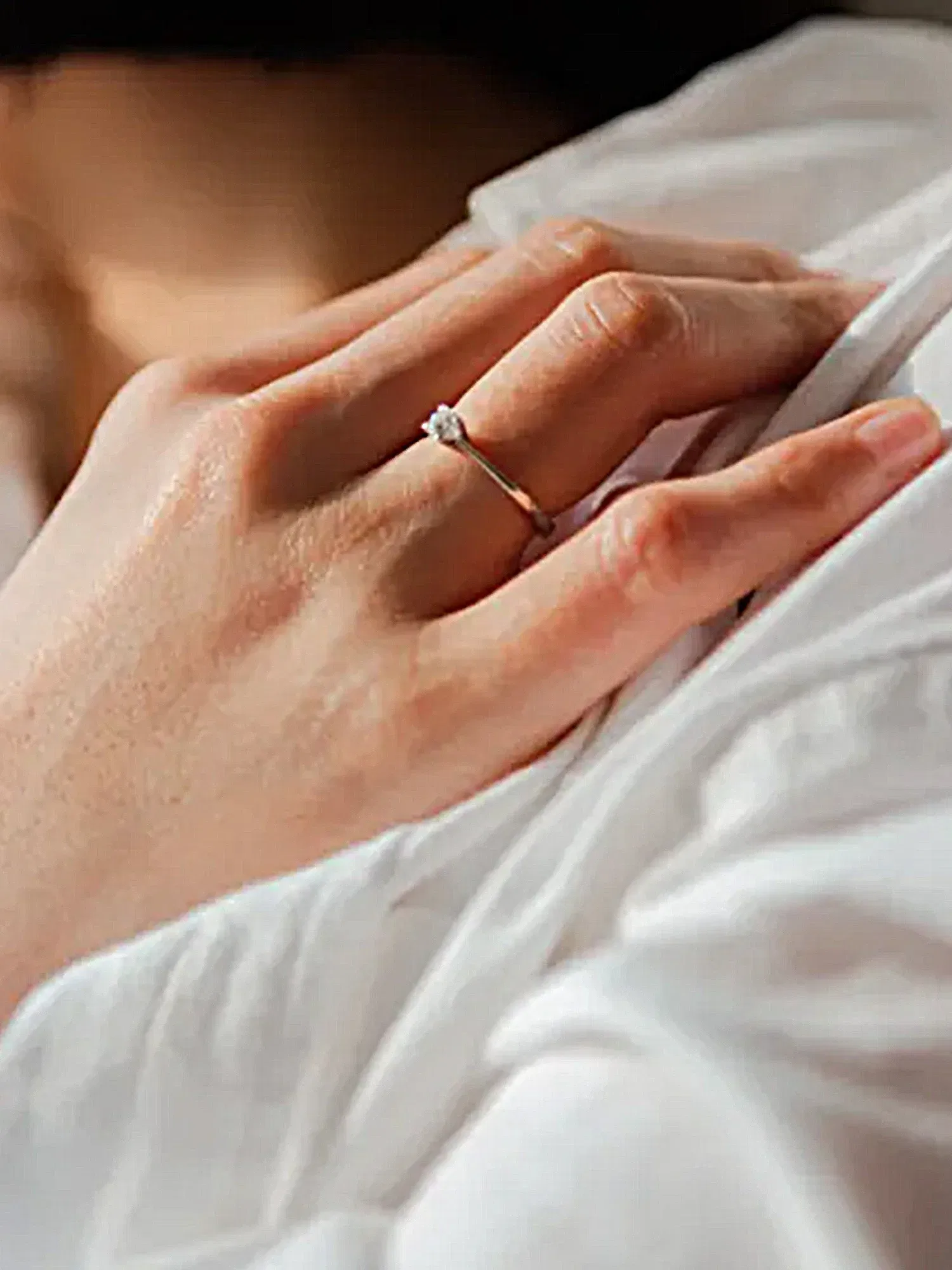 Eine Hand mit einem schlichten Verlobungsring liegt sanft auf weißem Stoff. Der silberne Ring ist mit einem Diamanten besetzt, der im Licht schimmert. Die Aufnahme konzentriert sich auf die Details des Rings, während die Hand sanft und entspannt erscheint.
