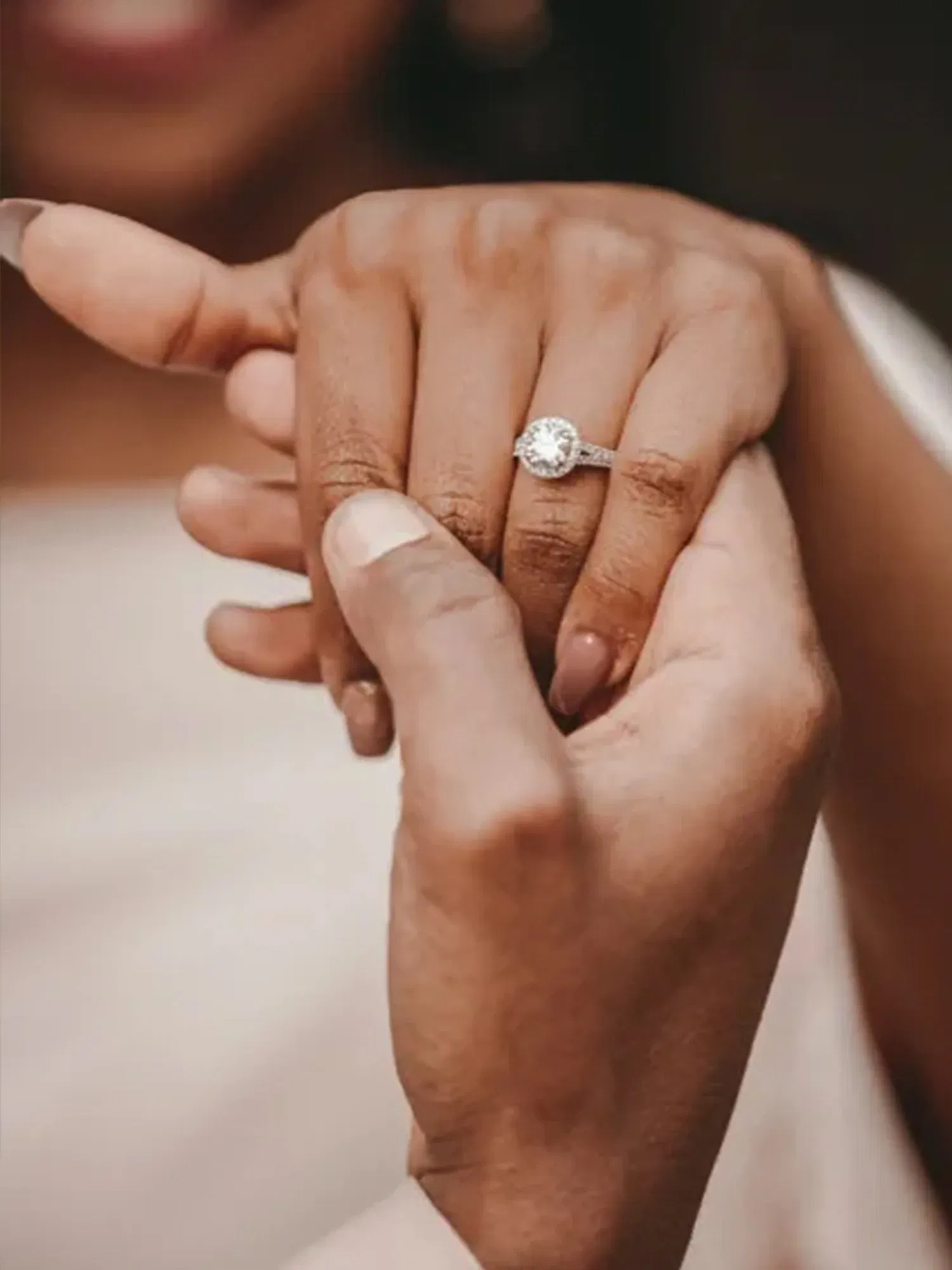 Eine Hand mit einem beeindruckenden Verlobungsring, gehalten von einer anderen Hand, beide Hände umschließen sanft den Ringfinger. Der Diamant des Rings glänzt im Licht, und im Hintergrund ist das verschwommene Gesicht der Frau zu sehen, die strahlend lächelt. Das Bild vermittelt eine intime Verlobungsszene.