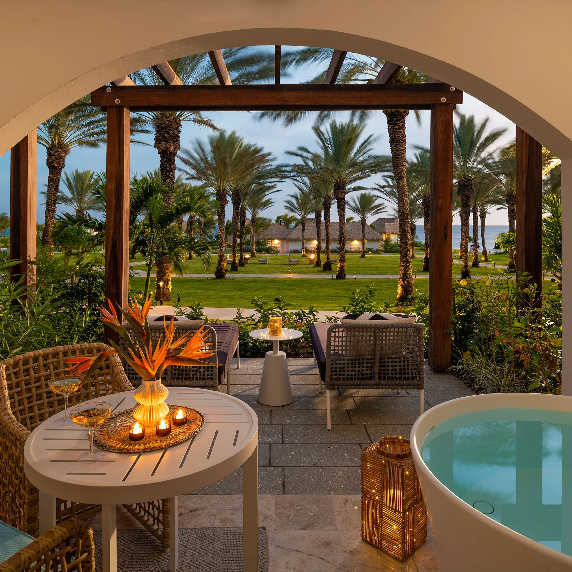 Eine tropische Veranda mit Ausblick auf Palmen und den Garten, dekoriert mit Kerzen und einer modernen, freistehenden Badewanne, unter einem offenen Bogen.