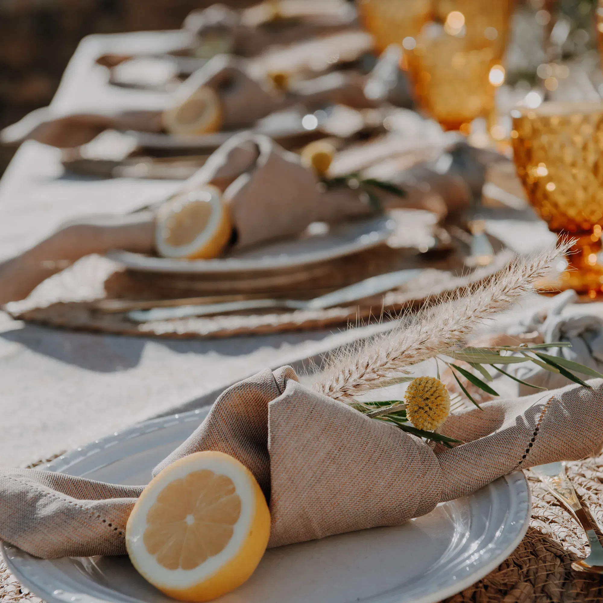 Detailaufnahme einer Hochzeitstafel mit natürlichen Dekorationselementen: Zitronen, Leinenservietten und goldenen Akzenten bei Sonnenuntergang.