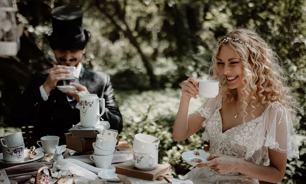 In einer verwunschenen Waldumgebung sitzen Braut und Bräutigam bei einer Teeparty, inspiriert von Alice im Wunderland, und genießen ihre Teetassen.