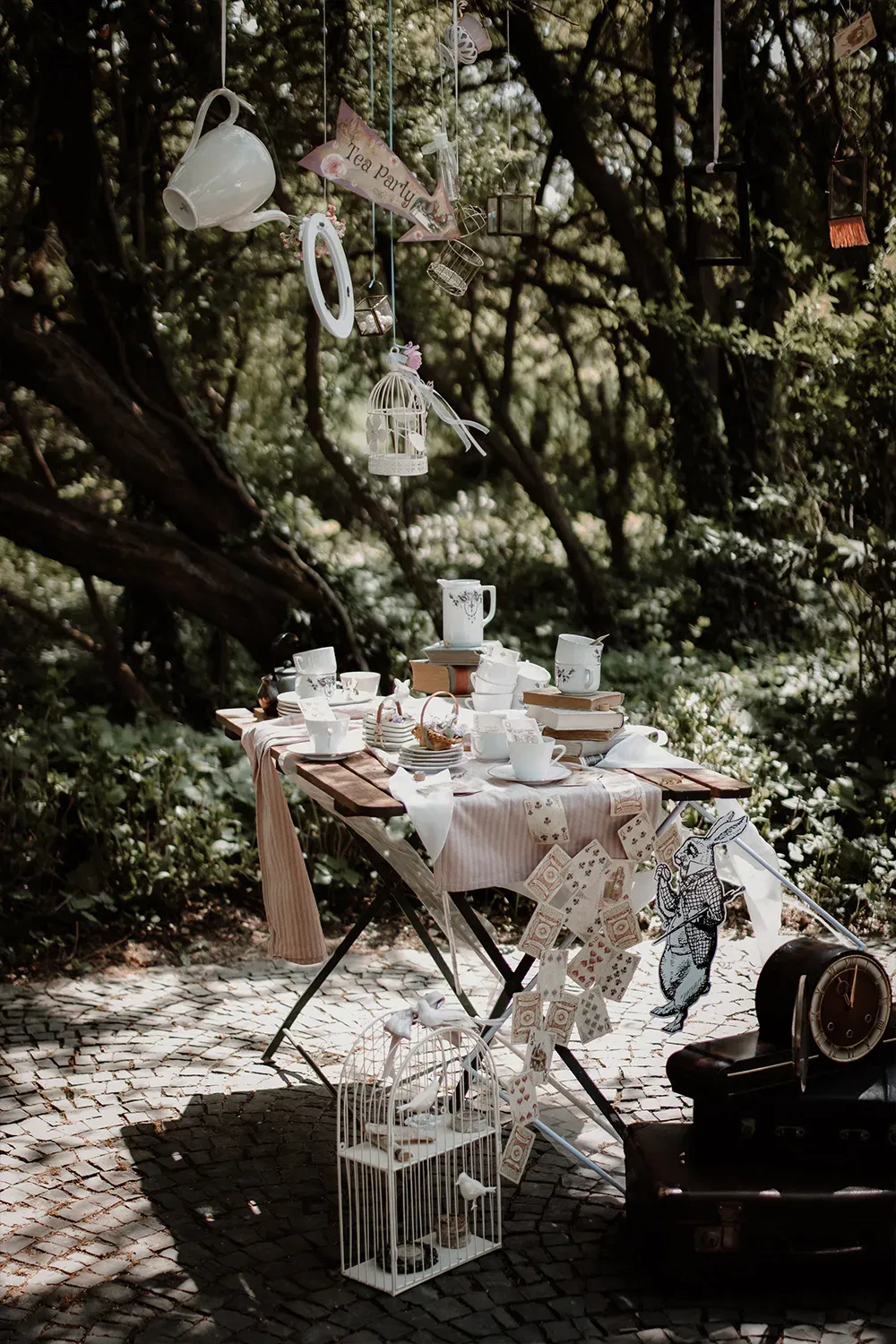Ein fantasievoll gestalteter Teetisch in einer Waldumgebung, geschmückt mit Tassen, Teller und schwebenden Teekannen, die von den Ästen hängen.