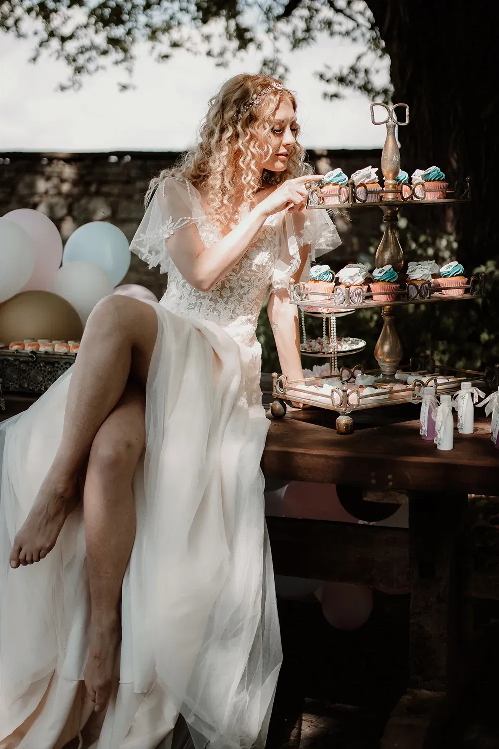 Braut im weißen Kleid, die Cupcakes von einem aufwendig dekorierten Ständer nimmt, in einem romantischen Garten-Setting mit Ballons im Hintergrund.
