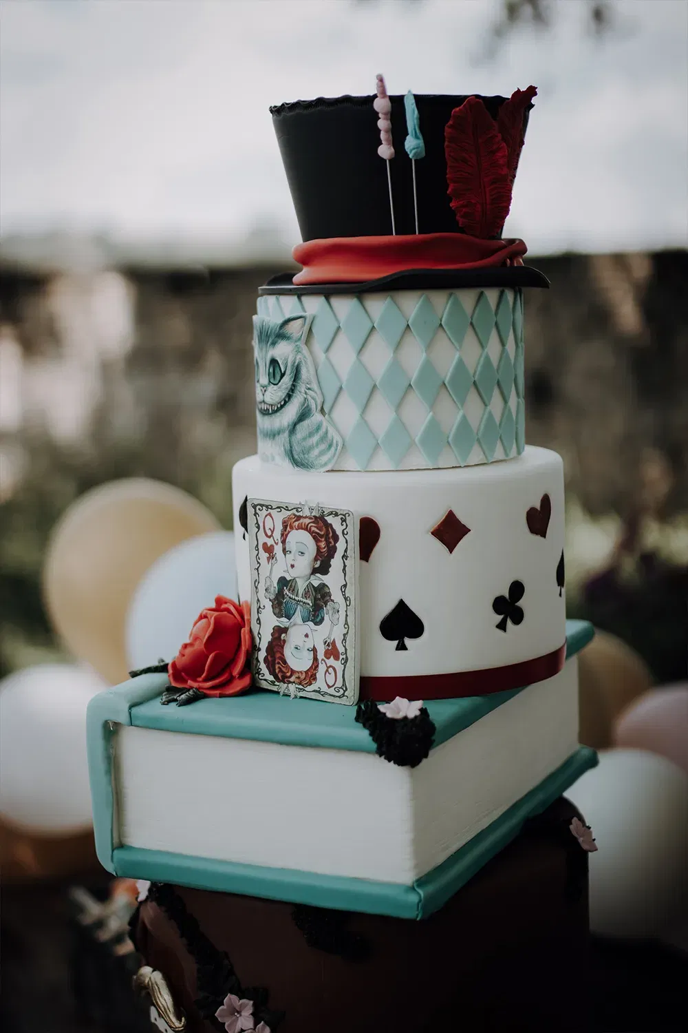 Kreative Hochzeitstorte im Fantasy-Stil, geschmückt mit Elementen aus "Alice im Wunderland", darunter ein Hut und Spielkarten als Dekoration.
