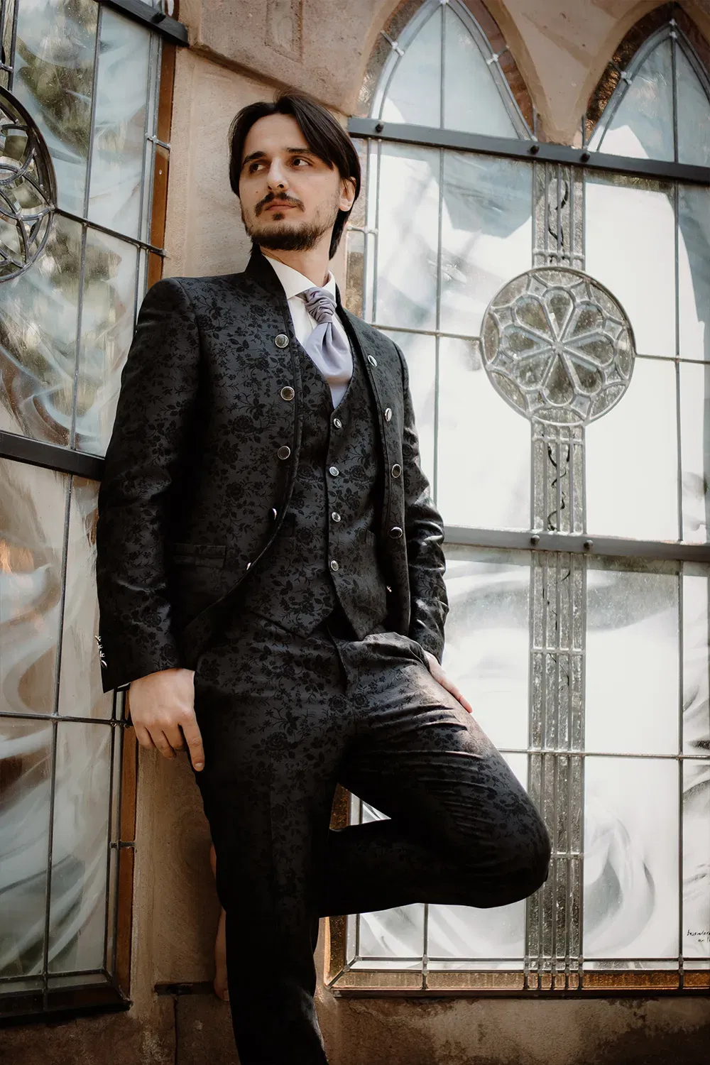 Der Bräutigam posiert stilvoll in einem gemusterten Anzug vor einem historischen Glasfenster, das Sonnenlicht fällt sanft durch die Fenster.