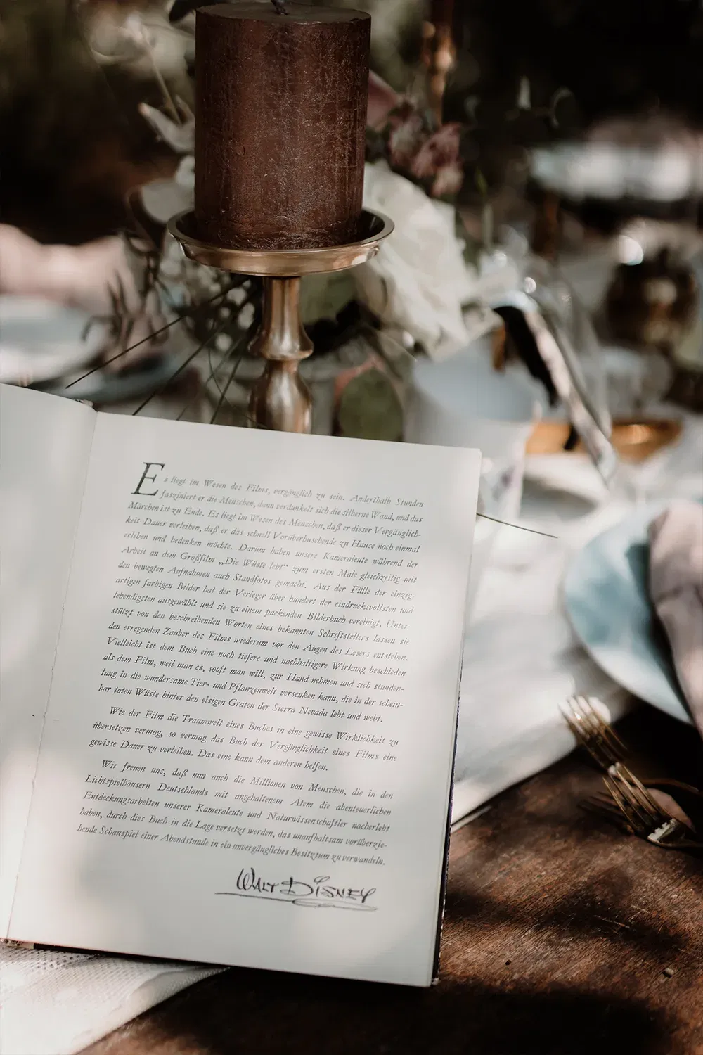 Auf einem elegant gedeckten Tisch ist ein Buch geöffnet, in dem ein inspirierendes Zitat von Walt Disney zu lesen ist, begleitet von Kerzenlicht.