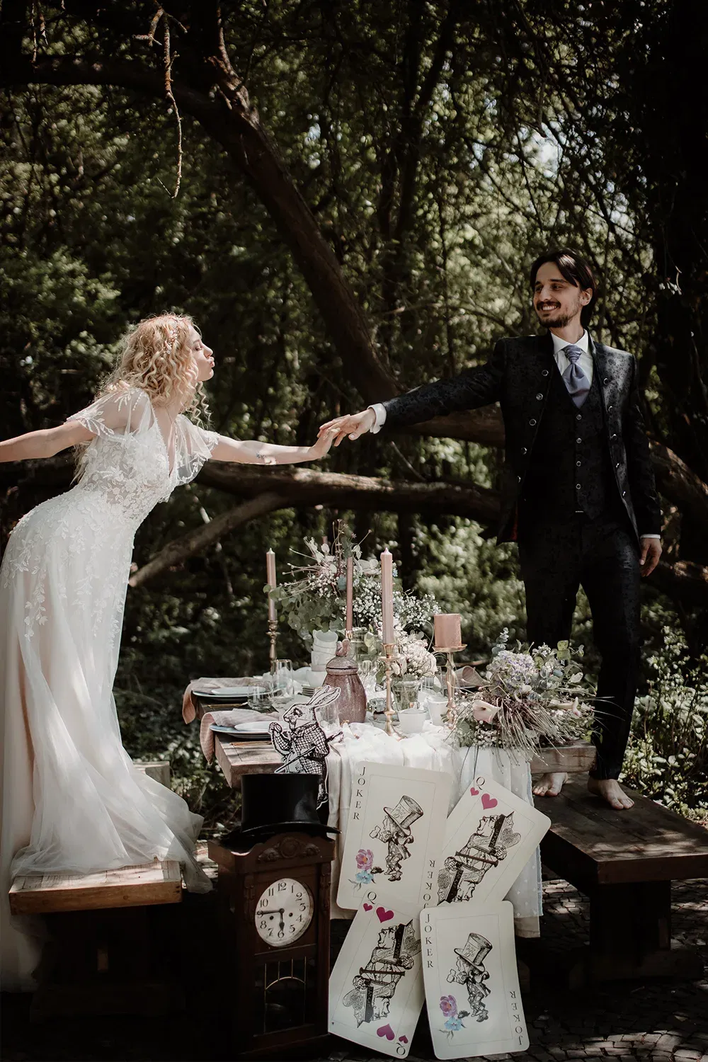 Das Brautpaar in eleganten Hochzeitskleidern posiert an einem fantasievoll gedeckten Tisch mit großen Joker-Spielkarten und einer alten Standuhr, während sie sich zärtlich die Hände reichen.