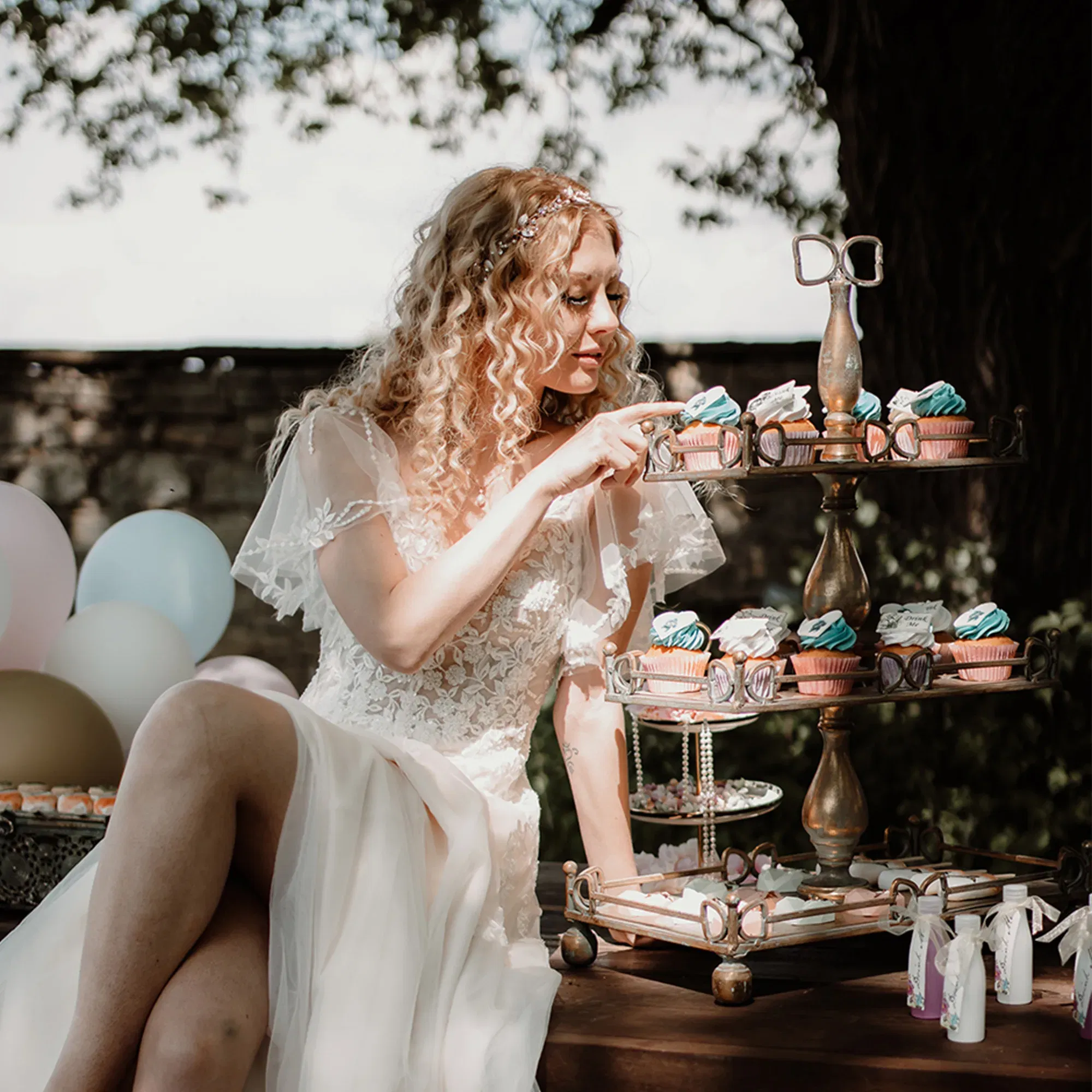 In einem weißen Hochzeitskleid sitzt die Braut neben einer Etagere voller Cupcakes und wählt vorsichtig einen aus. Bunte Luftballons ergänzen die verträumte Atmosphäre der Szene.