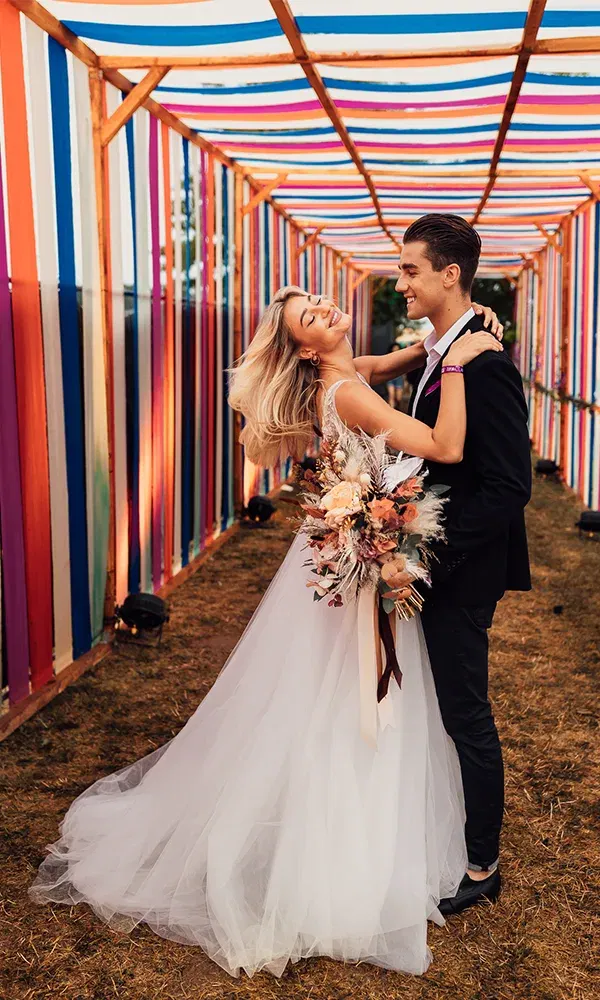 Unter einem überdachten, regenbogenfarbenen Baldachin tanzen Braut und Bräutigam in einem fröhlichen Moment der Zweisamkeit. Der Bräutigam umarmt die Braut zärtlich, die mit einem strahlenden Lächeln ihren Blumenstrauß präsentiert.