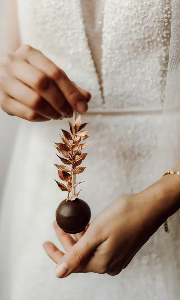 Nahaufnahme eines dekorativen Cake-Pops mit goldener Verzierung, der von einer Braut in einem perlenbestickten Kleid präsentiert wird.