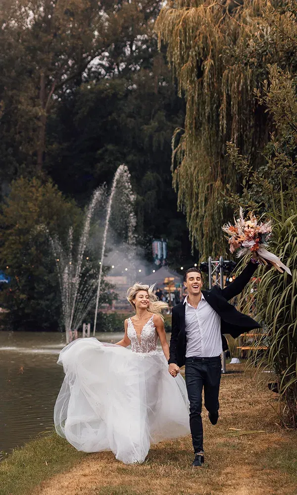 In einem natürlichen, parkähnlichen Setting rennt das Hochzeitspaar lachend am Ufer eines Sees vorbei. Die Braut trägt ein üppiges weißes Kleid, während der Bräutigam fröhlich den Blumenstrauß schwenkt, begleitet von romantischen Wasserspielen.