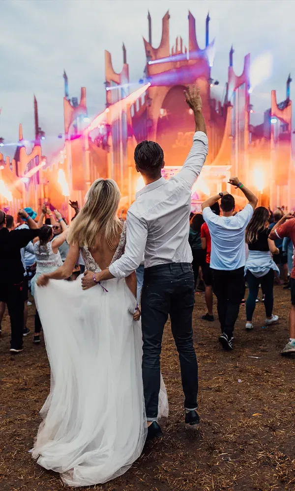 Das Hochzeitspaar steht auf einem Festivalgelände vor einer Bühne, umgeben von Menschen. Die Braut trägt ein elegantes weißes Kleid, der Bräutigam hebt jubelnd eine Hand.