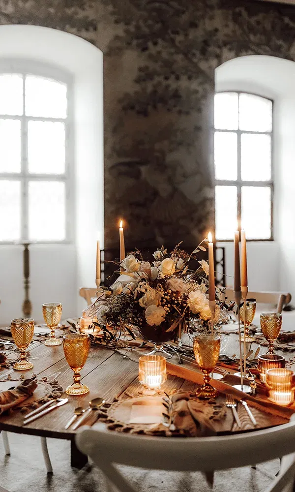 Ein festlich geschmückter Tisch mit floralen Details, goldenen Gläsern und warmem Kerzenschein, arrangiert in einem historischen Raum.