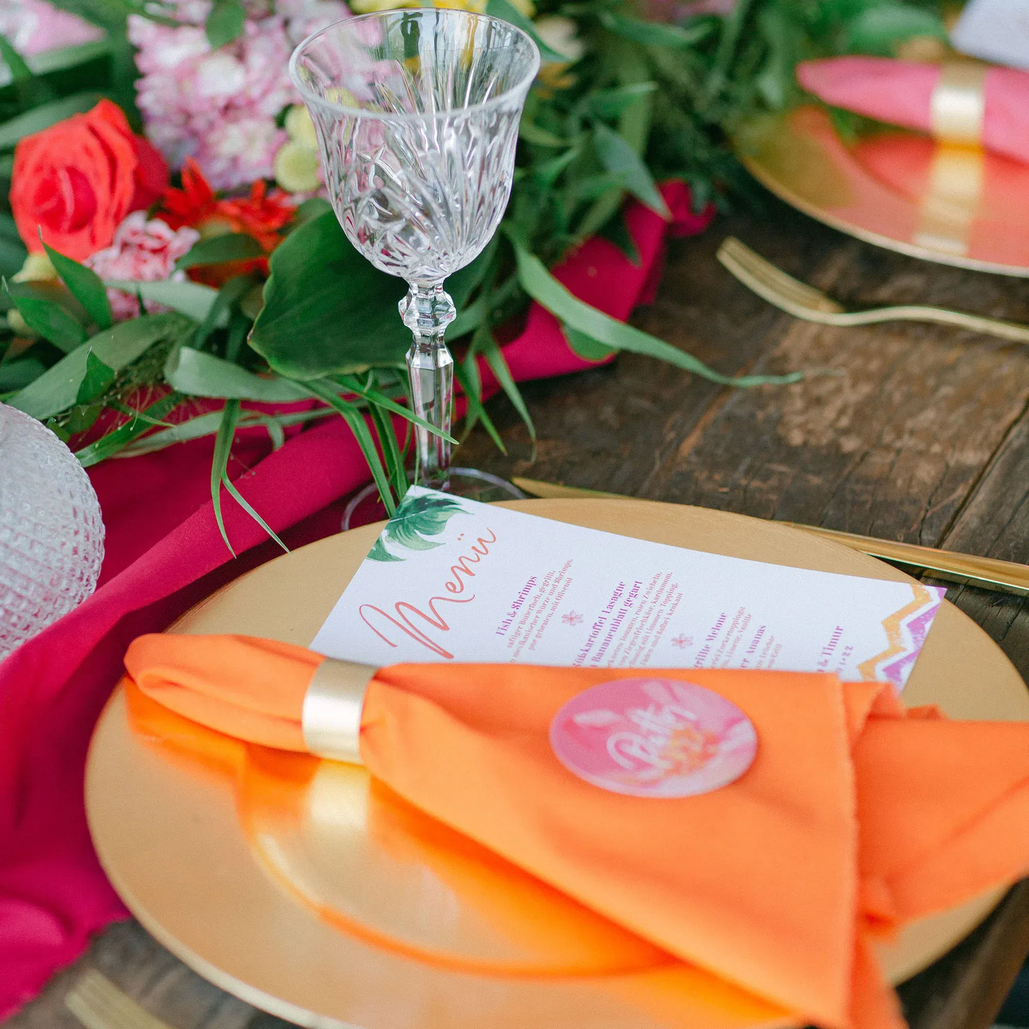 Eine stilvolle Tischdekoration mit einer orangefarbenen Serviette, die mit einem goldenen Serviettenring auf einem goldenen Teller liegt. Daneben befindet sich ein Kristallglas und ein farbenfrohes Menü auf dem Tisch.