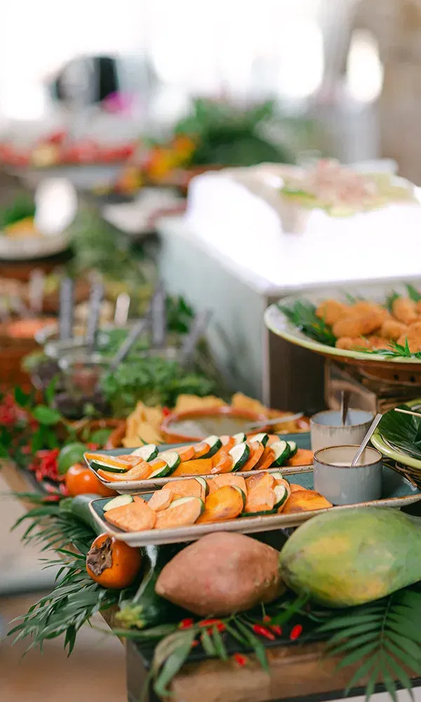 Ein Hochzeitsempfangsbuffet mit frischen, saisonalen Gerichten. Zucchini und Süßkartoffeln liegen auf einem stilvollen Tablett, während tropische Früchte und dekorative Blätter das Arrangement abrunden.
