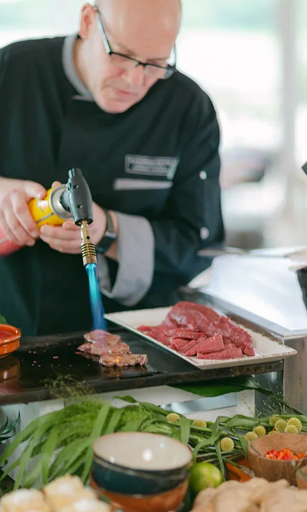 Professioneller Koch benutzt einen Flambierbrenner, um Fleisch auf einer heißen Platte zu garen, während verschiedene grüne Zutaten auf dem Tisch liegen.