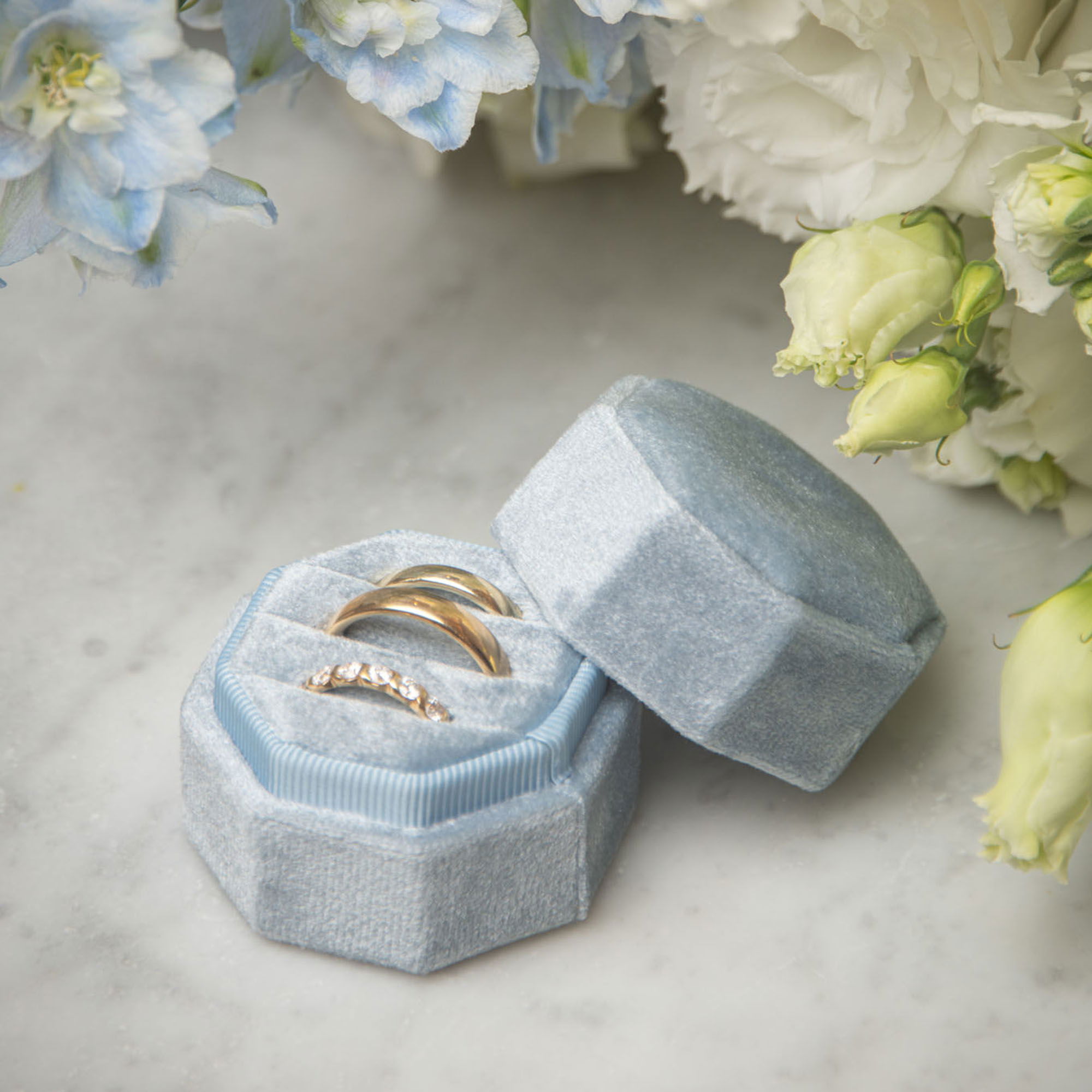 Goldene Eheringe in einem pastellblauen Samtkästchen, dekoriert mit weißen Rosen und blauen Blüten auf einer hellen Marmorplatte.