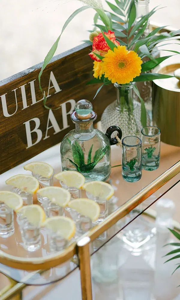 Eine einladende Tequila-Bar mit kunstvoll gestalteten Shotgläsern, Zitronenscheiben und dekorativen Blumen, die das tropische Thema der Feier unterstreichen.