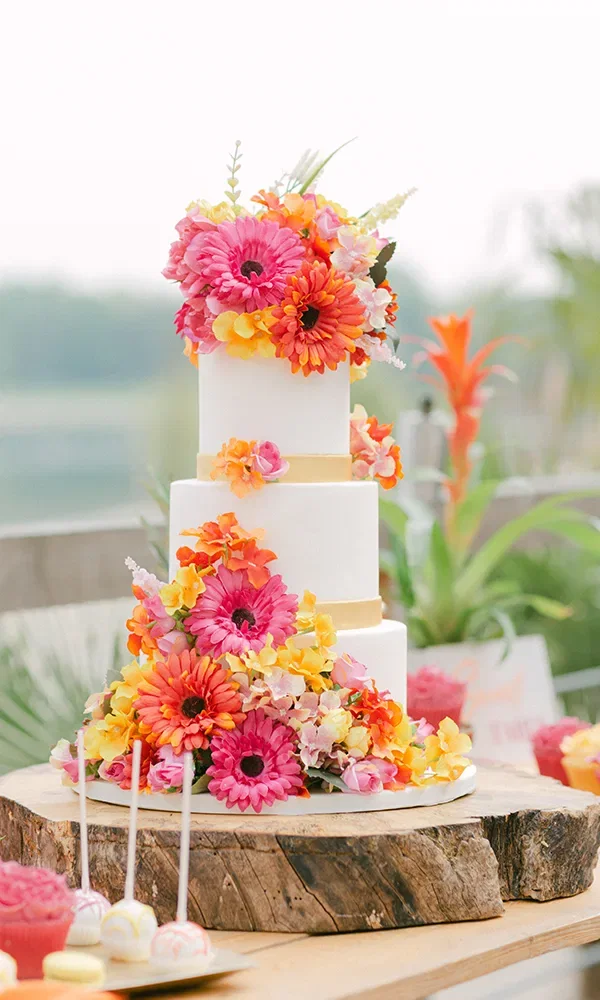 Eine schicke Hochzeitstorte mit mehreren Schichten, dekoriert mit leuchtenden Blüten in Rosa und Gelb. Sie steht auf einem Holzteller und ist das Highlight der sommerlichen Hochzeitsdekoration.
