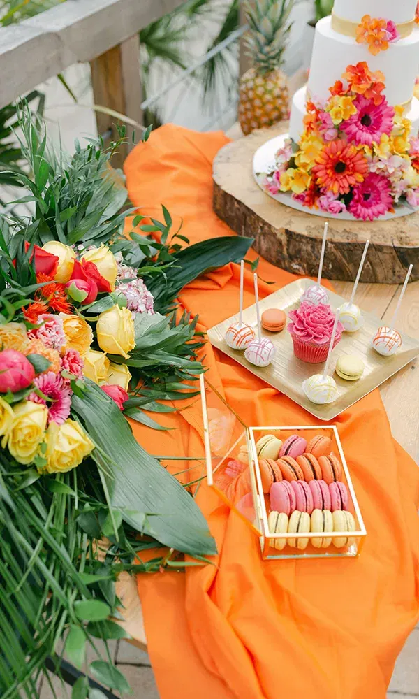 Eine tropisch dekorierte Hochzeitstorte mit bunten Blumen, umgeben von Macarons und Cupcakes, ist auf einem festlich dekorierten Tisch mit leuchtenden Blüten und grünen Blättern zu sehen.