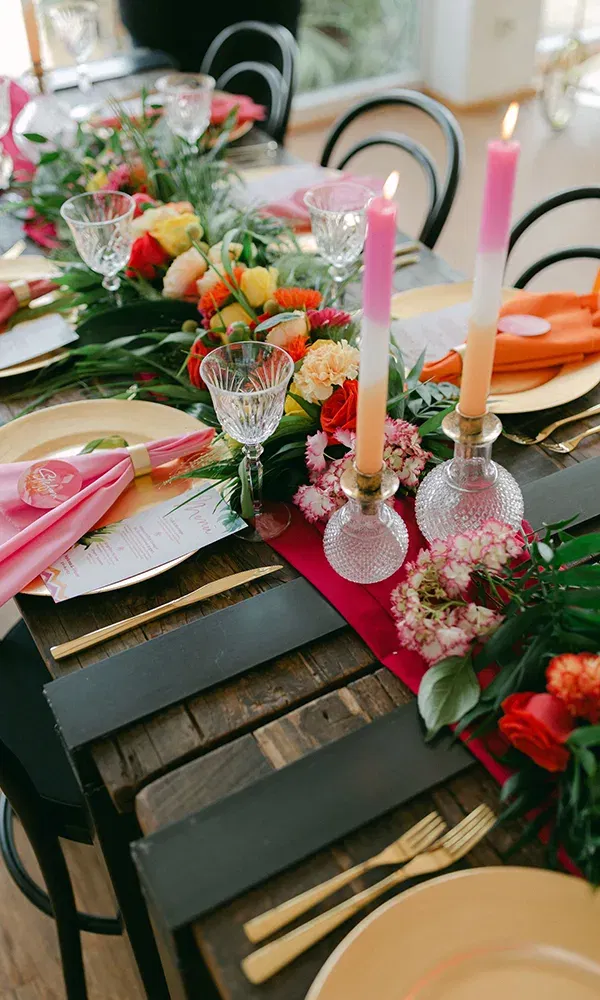 Eine farbenfrohe Tischdekoration mit rosa und orangefarbenen Servietten, Kristallgläsern und leuchtenden Kerzen in Vasen, die auf einem Holztisch arrangiert sind. Frische Blumen verleihen der Szene lebendige Farben.