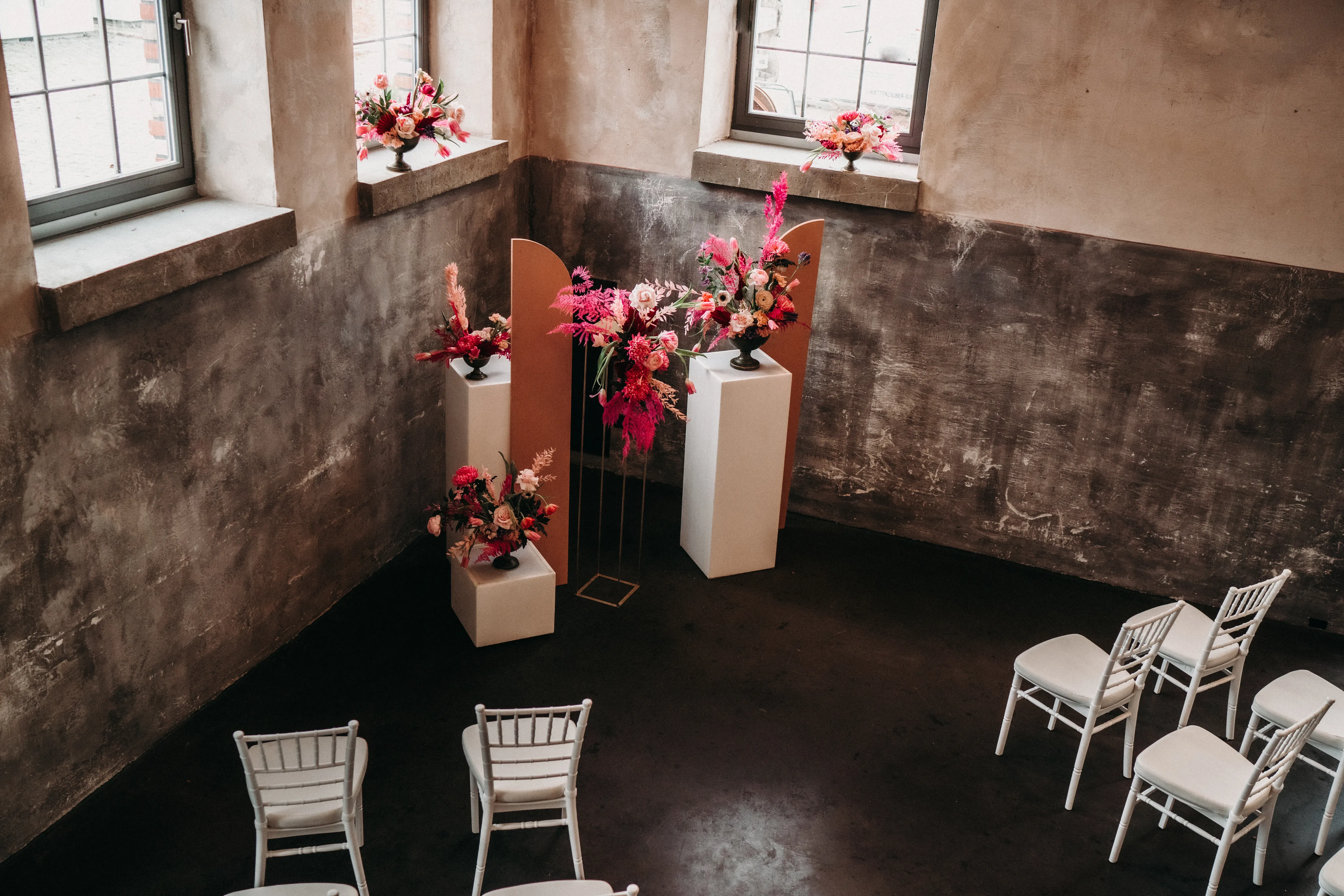Ein stilvoller Raum für Zeremonien mit schlichten, hellen Wänden und großen Fenstern, durch die Tageslicht einfällt. In der Mitte des Raumes befinden sich Blumendekorationen in kräftigen Pink- und Rottönen, die auf Podesten stehen, während weiße Stühle in einer offenen Formation um den Bereich angeordnet sind.