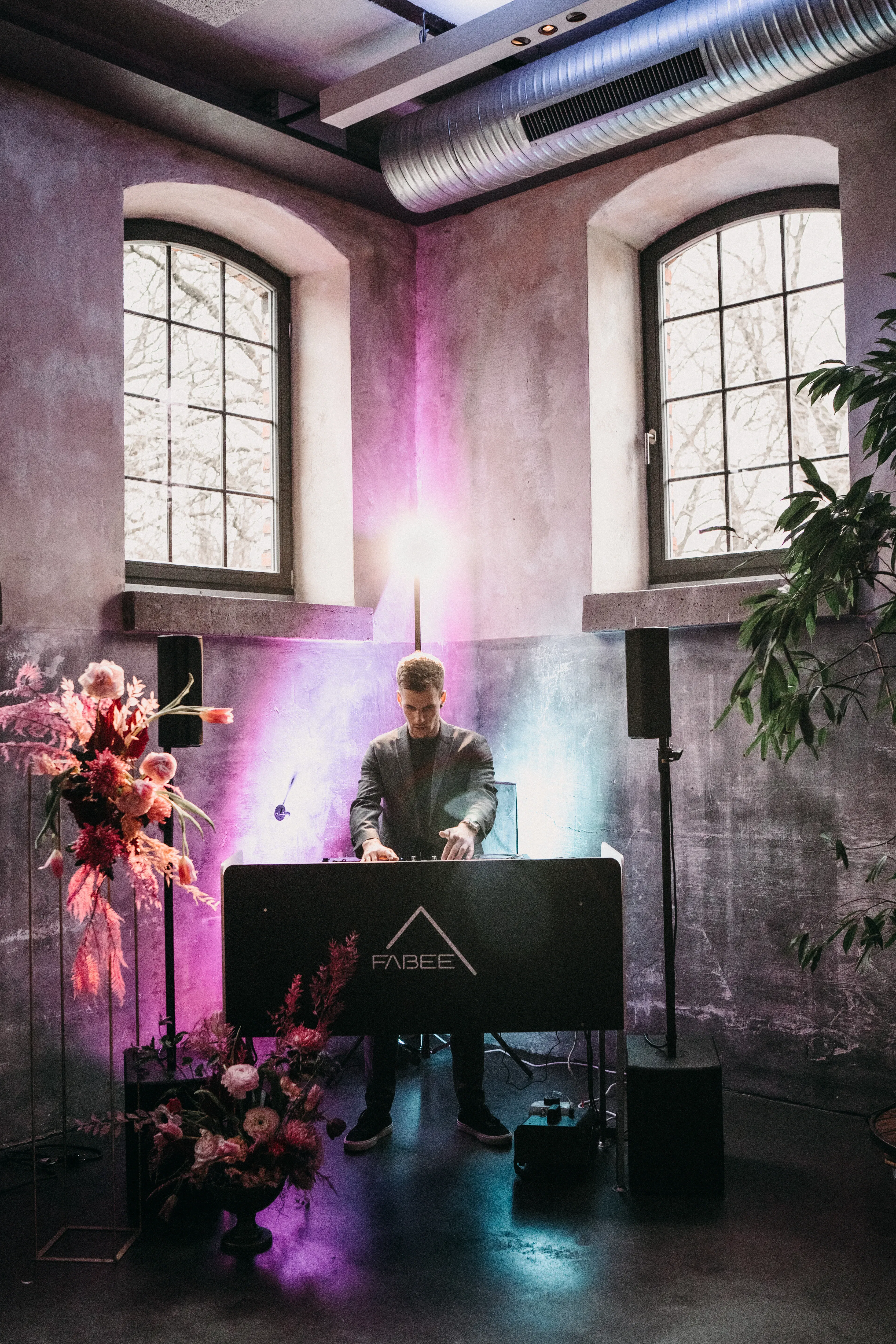 Der DJ ist in einem industriell gestalteten Raum mit großen Fenstern und modernen Lichteffekten in den Farben Lila und Blau zu sehen. Neben ihm befinden sich Blumenarrangements in lebhaften Farben, die den sonst schlichten Raum auflockern. Der DJ konzentriert sich auf sein Set an einem schwarzen Pult.