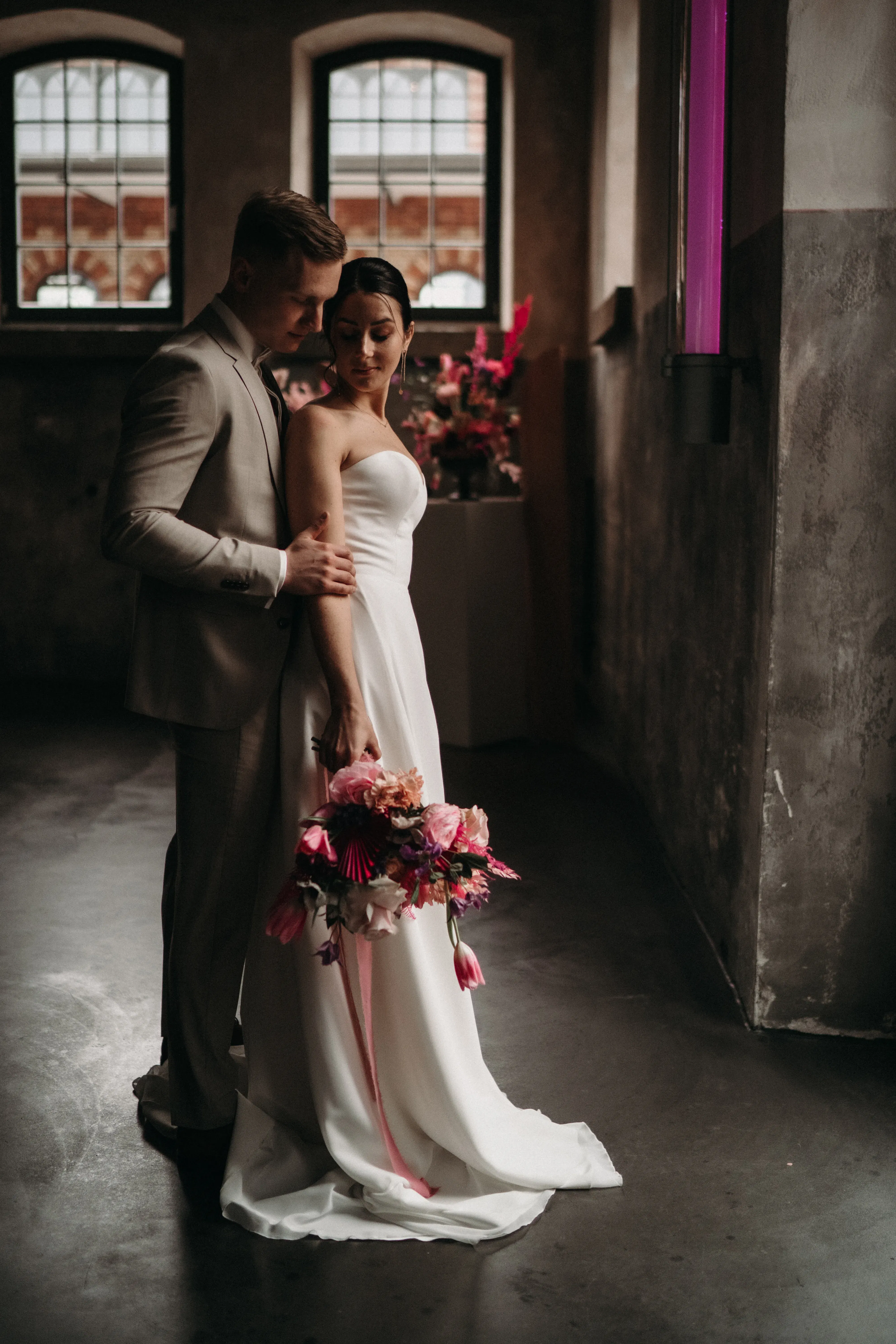 Eine romantische Szene mit einem Hochzeitspaar in einem stilvollen Raum mit hohen Fenstern. Die Braut, in einem schlichten Kleid mit Blumenstrauß, lehnt sich liebevoll an den Bräutigam, der sie zärtlich hält. Ihr bunter Blumenstrauß in kräftigem Rosa und Pink bildet einen schönen Kontrast zum schlichten Ambiente.
