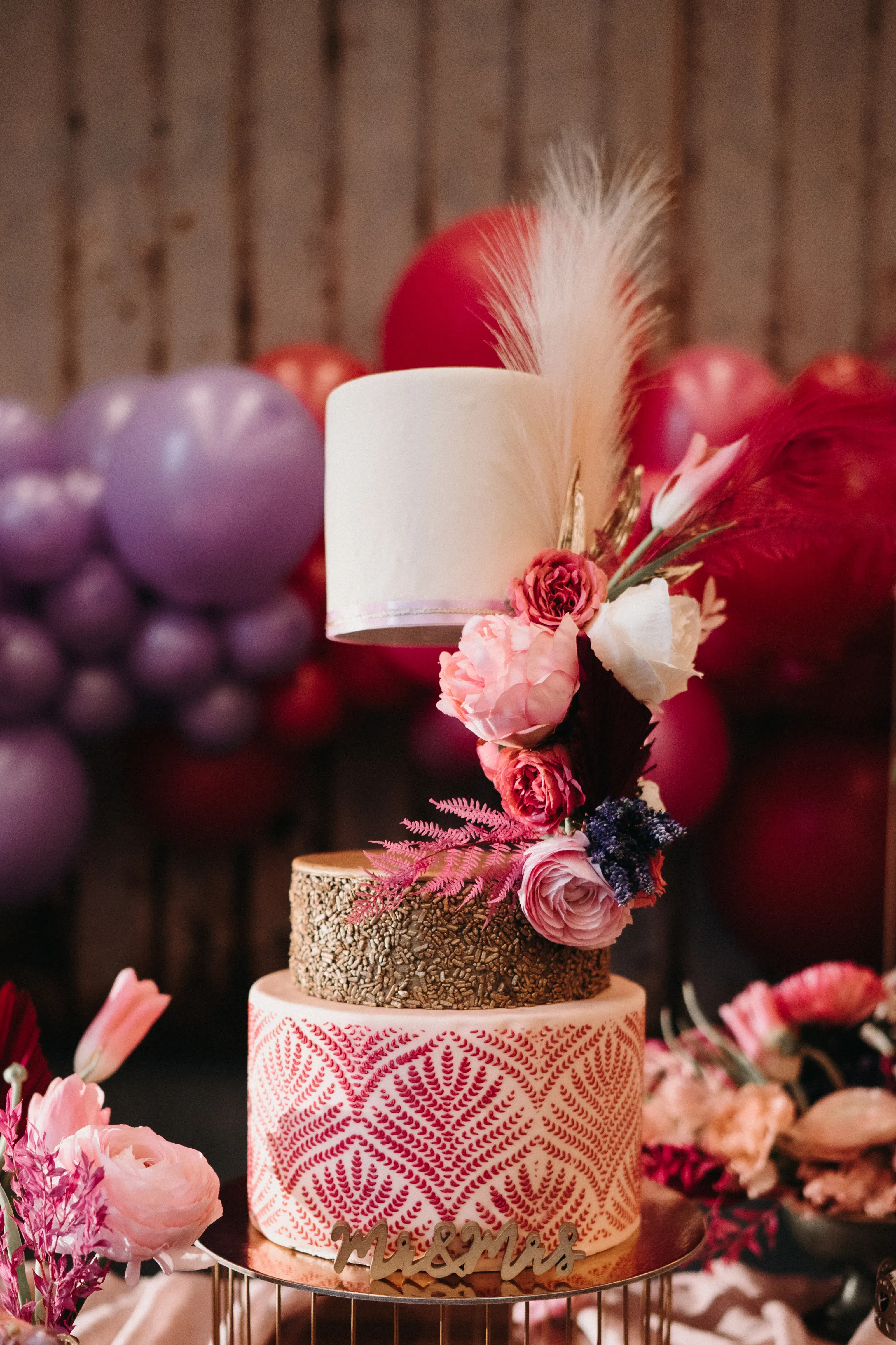 Eine farbenfrohe Hochzeitstorte mit drei Ebenen, dekoriert mit pinken und roten Blüten. Die Torte hat eine weiße Spitze, eine goldene Mitte und eine untere Schicht mit einem geometrischen Muster in Pink. Hinter der Torte sieht man eine Kulisse aus lilafarbenen und roten Ballons, die eine festliche Atmosphäre schaffen.