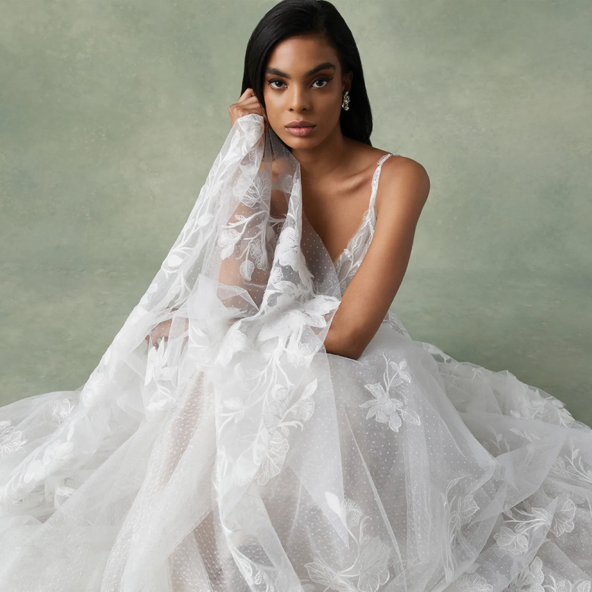 Braut in einem märchenhaften, weißen Kleid mit aufwändigen Spitzenverzierungen, die den Schleier um ihre Schultern drapiert, sitzt vor einem minimalistischen, grünlichen Hintergrund und verleiht dem Bild eine elegante Atmosphäre.