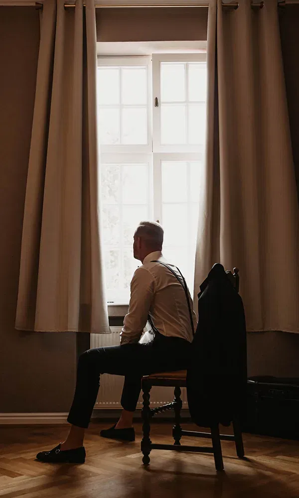 Ein Bräutigam sitzt still vor einem Fenster, durch das Tageslicht strömt. Der Raum ist in warmes Licht getaucht, und er trägt elegante Kleidung mit Hosenträgern. Sein Jackett hängt über dem Stuhl.