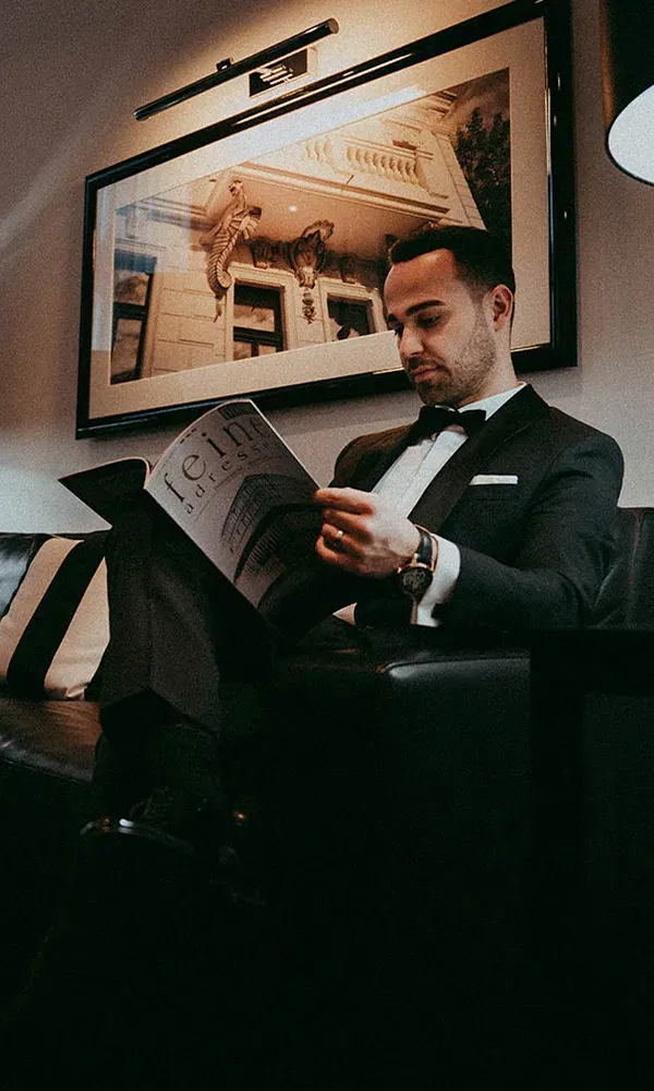 Der Bräutigam sitzt ruhig auf einem Sofa und liest in einem Magazin, während eine stilvolle Lampe neben ihm steht. Hinter ihm hängt ein gerahmtes Bild, das einen prächtigen Balkon zeigt.