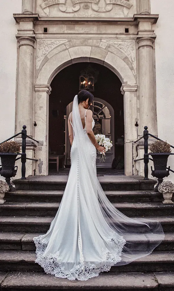 Die Braut, in einem langen weißen Kleid und Schleier, steht auf einer Treppe vor einem alten, kunstvoll verzierten Eingang. Der Rücken ist der Kamera zugewandt, das Kleid und der Schleier fließen elegant über die Stufen. Sie trägt einen Blumenstrauß.