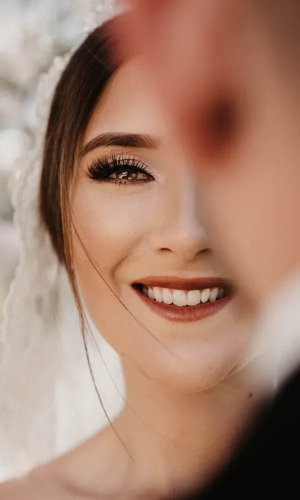 Detaillierte Nahaufnahme einer Braut, die sanft lächelt. Ihr Gesicht ist leicht von einer verschwommenen Person verdeckt. Die Braut hat lange Wimpern, dezentes Make-up und einen Schleier, der sanft ihr Haar umrahmt.