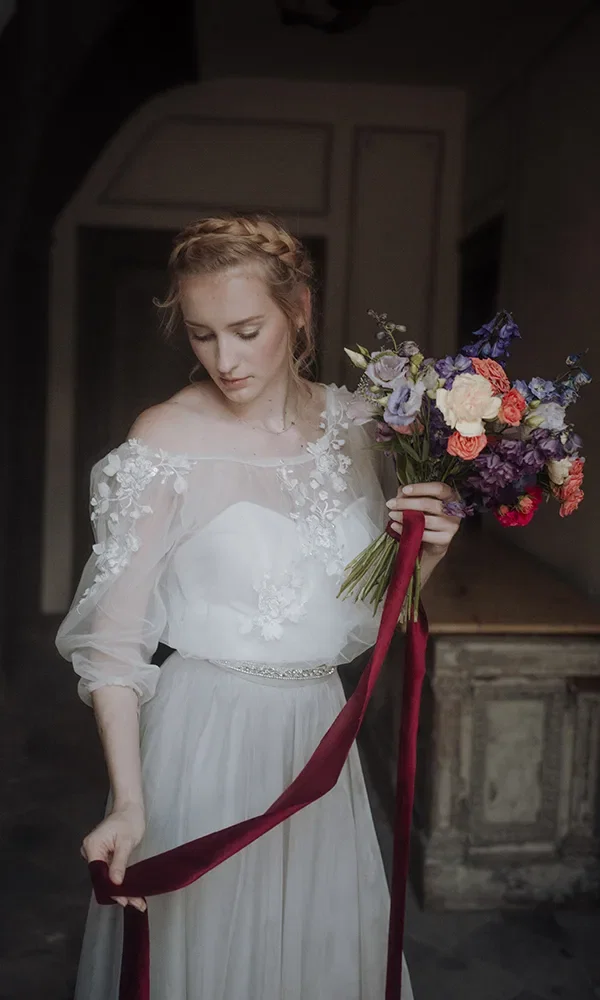 Die Braut, die ein weißes Kleid mit Blumenapplikationen trägt, hält einen Strauß bunter Blumen mit einem langen roten Band. Sie blickt sanft nach unten, während das Band in ihrer Hand fällt.