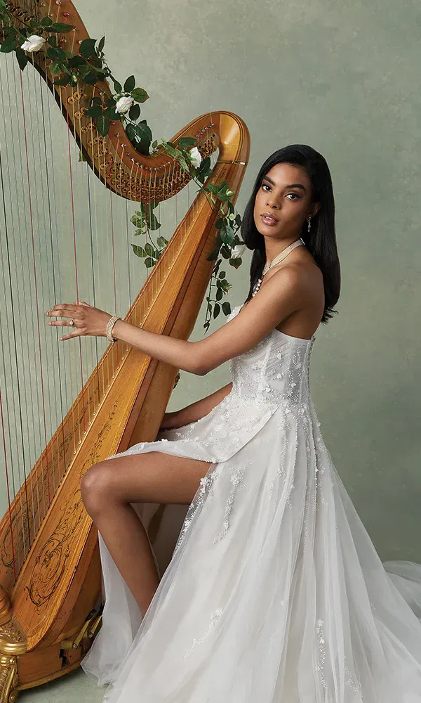 Eine stilvolle Braut sitzt in einem perlenbesetzten Hochzeitskleid neben einer großen, kunstvoll dekorierten Harfe. Das lange, weiße Kleid fließt zu Boden, und die Braut strahlt eine elegante Ruhe aus, während ihr Gesichtsausdruck selbstbewusst und weich wirkt. Die Harfe ist mit Blumen geschmückt und verstärkt die romantische Atmosphäre.