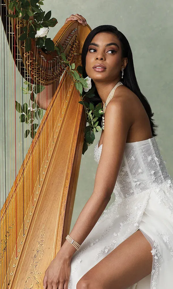 Die Braut posiert an einer Harfe, umgeben von floralen Akzenten. Ihr langes Haar fällt sanft auf ihre Schultern, und das weiße, mit Perlen verzierte Kleid ergänzt den warmen Goldton der Harfe. Sie wirkt ruhig und anmutig, während sie die Harfe leicht berührt.