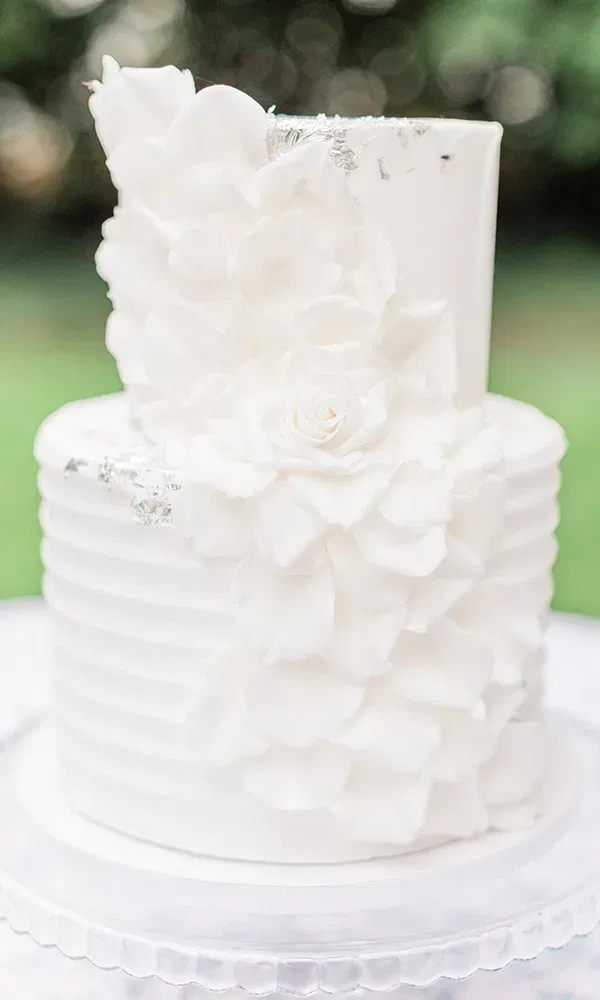 Eine schlichte, aber elegante Hochzeitstorte mit weißen Blüten und silbernen Details steht im Freien auf einem gläsernen Tortenständer, während die grüne Natur den Hintergrund bildet.