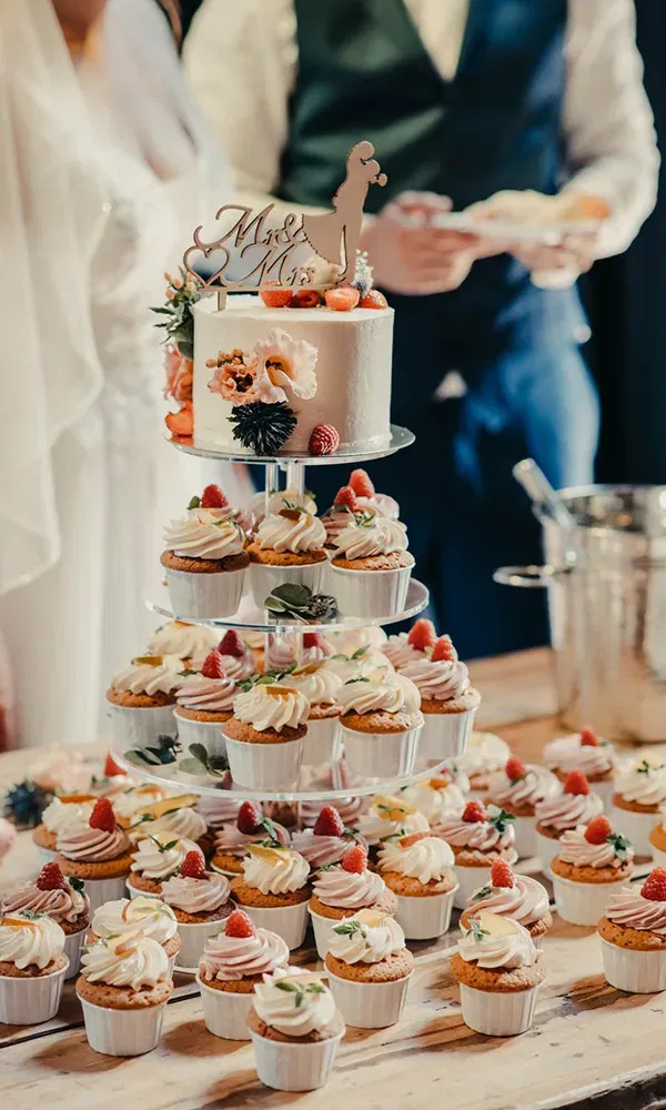 Die Hochzeitstorte und die dazugehörigen Cupcakes auf einer Etagere sind kunstvoll mit frischen Früchten dekoriert. Die Torte trägt eine "Mr. & Mrs."-Topperfigur, und das Brautpaar im Hintergrund bereitet sich darauf vor, die Torte anzuschneiden.