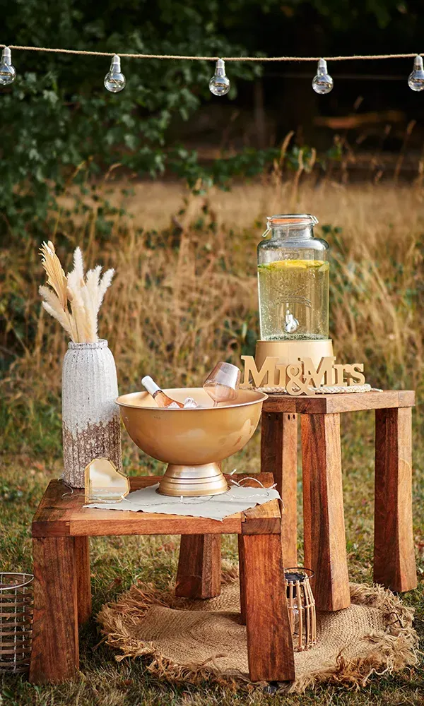 Eine stilvolle Getränkestation auf einem rustikalen Holztisch mit einer goldenen Schale voller Eis und Getränken. Daneben ein Getränkespender mit Zitronenwasser und eine Dekoration mit Trockenblumen sowie einer "Mr & Mrs"-Holzschrift.