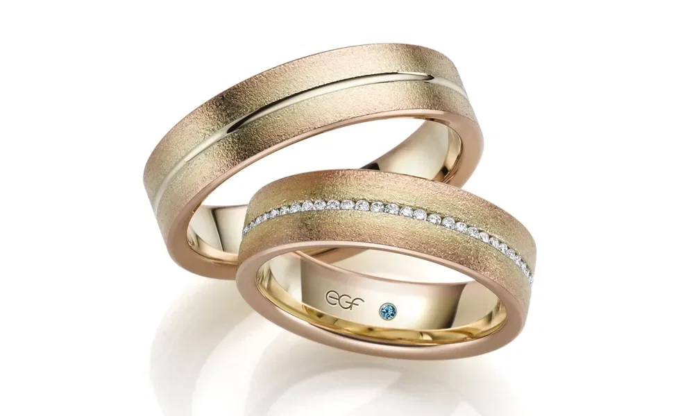 Ein Paar modern gestalteter Trauringe in warmen Goldtönen. Der Ring der Braut hat eine Reihe kleiner Diamanten, die entlang der Mitte funkeln, während der Ring des Bräutigams eine dezente Vertiefung entlang der Oberfläche zeigt. Beide Ringe haben eine matte, gebürstete Textur und schimmern leicht.