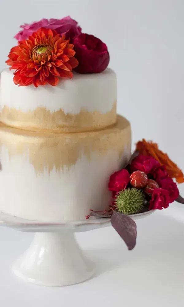 Eine stilvolle zweistöckige Torte, verziert mit goldenen Schattierungen und leuchtenden Blumen, steht auf einem eleganten, weißen Tortenständer und sorgt für einen bunten Akzent auf der Hochzeitstafel.