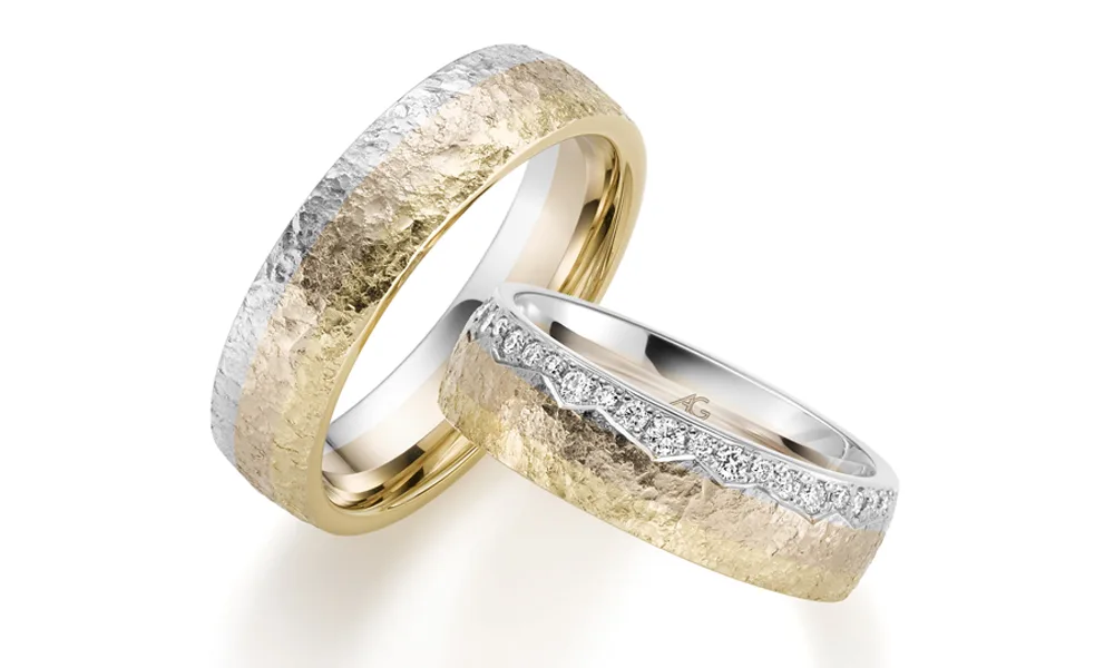 Ein Set aus zwei Eheringen mit einer markanten Struktur, bei dem Gelb- und Weißgold nahtlos ineinander übergehen. Der Damenring ist mit einer Reihe funkelnder Diamanten entlang des oberen Randes geschmückt, während der Herrenring eine gehämmerte, matte Textur aufweist.