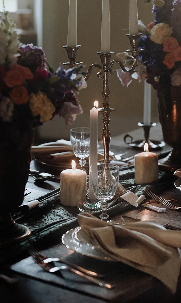 Eine stilvolle Tischdekoration mit Kerzen, Blumen und silbernen Kerzenständern. Zwischen den brennenden Kerzen sind Gläser, Teller und Servietten auf einem Holztisch kunstvoll arrangiert.
